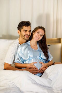 Romantic Caucasian husband tenderly embrace lovely pregnant wife on bed under quilt and carefully touch belly as expecting for birth of unborn baby