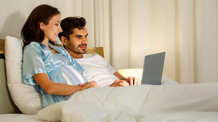 Fototapeta premium Cheerful Caucasian pregnant woman smiling as enjoy use laptop computer for health nourishment to prepare birth of unborn baby while resting on bed in bedroom of condominium.
