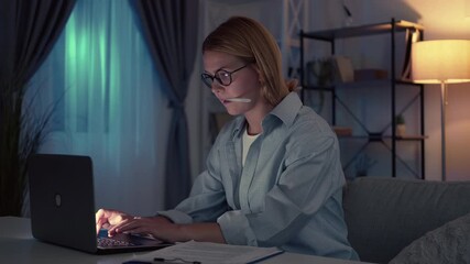 Late work. Busy woman. Online research. Concentrated casual lady working hard on laptop making notes sitting in dark light room interior.