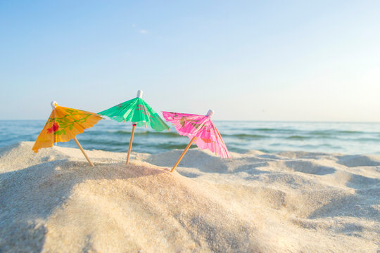 Three paper cocktail multi-colored umbrellas on sandy sea beach with blue sky on sunny summer day close-up. Concept Festive Celebrating Party New Year travelling vacation tourism rest relax holiday