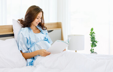 Cheerful Caucasian pregnant woman smiling as enjoy reading book for health nourishment to prepare birth of unborn baby while resting on bed in bedroom of condominium.