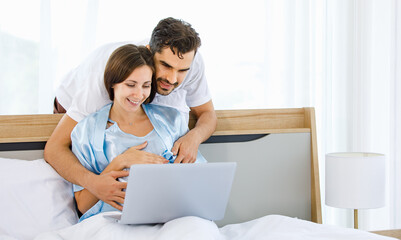 Cheerful Caucasian pregnant woman resting on bed and husband embrace from back to tenderly touch...