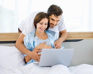 Cheerful Caucasian pregnant woman resting on bed and husband embrace from back to tenderly touch belly of unborn baby with love and care