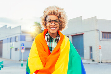 Cheerful smiling black woman with eyeglasses, headsets, afro hair, rainbow flag of gay pride on a sunny day outdoors. She fights for sexual freedom. - obrazy, fototapety, plakaty