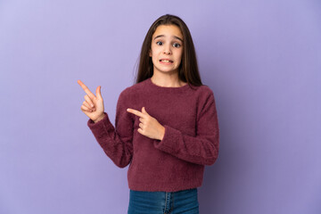 Little girl isolated on purple background frightened and pointing to the side