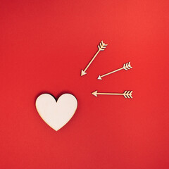 Natural wooden heart shape is targeted by arrows on bold red background. Minimal ready for romance concept with copy space. Flat lay Cupid love idea. Valentines day Holiday.