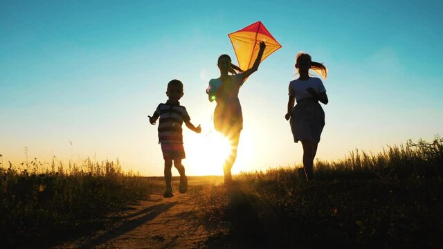 Happy young mother with a children play kites together on park. Boy running with flying kite in hand. Silhouette of happy people running at sunset in the park. Happy parents childhood dream concept.