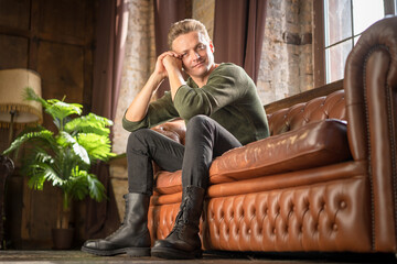 Mature Macho Man in Black Ankle Boots Looks Down at the Camera and Sits on a Brown Leather Sofa in...