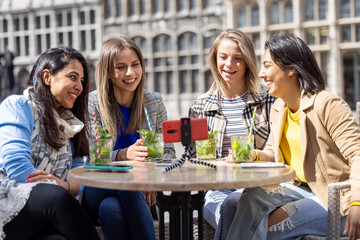 Antwerp, Belgium, May 21, 2021, four mixed race female tourists or students sitting outside in the old city center at a cafe terrace holding a video call using mobile phone. High quality photo