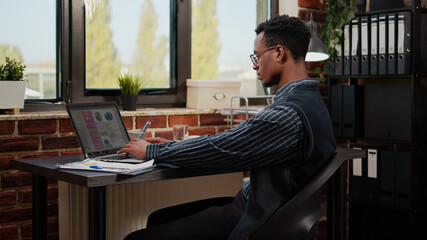 African american person working on e commerce statistics in company office. Business man using laptop to plan commercial report and financial investment for manager career development.