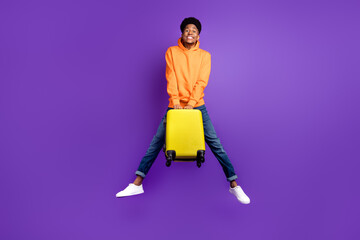 Full size photo of young black man happy positive smile jump hold suitcase travel vacation isolated over violet color background - obrazy, fototapety, plakaty