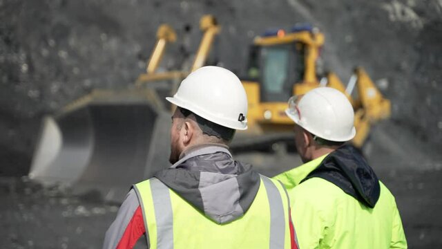 Two miners in special clothes and helmets stand in front of a passing bulldozer.
