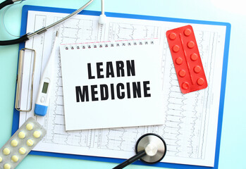 A notepad with the text LEARN MEDICINE lies on a medical clipboard with a stethoscope and pills on...