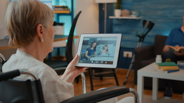 Elder person in wheelchair holding tablet with online video call to chat with family in hospital. Retired adult with physical disability talking on teleconference for remote communication.