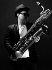 Black and White. Young Saxophonist in a Black Jacket, Hat and Sunglasses Holds a Baritone...