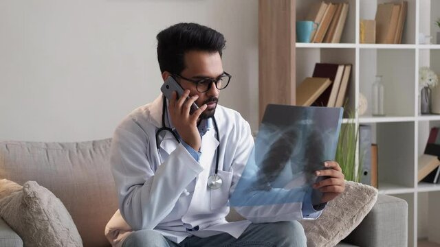 Online consultation. Male doctor. Labs result. Smart man talking with patient mobile phone looking x-ray photo of lungs in light clinical room interior.