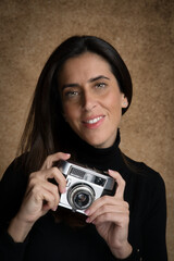 Retrato de joven mujer atractiva sonriente con cámara de fotos retro, con fondo desenfocado marrón y jersey negro.