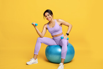 Fototapeta na wymiar Happy black lady in sports clothes sitting on fitball and working out with dumbbells on yellow studio background
