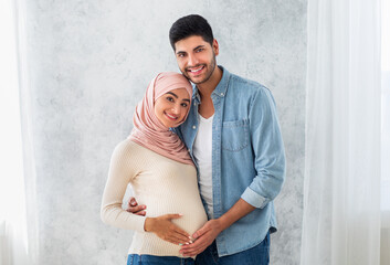 Portrait of young pregnant arab spouses, embracing and smiling at camera, loving muslim man...