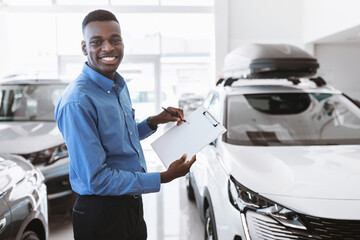 Cheerful black car salesman holding empty clipboard, smiling at camera in modern auto dealership,...