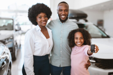 Cute black girl showing car key, standing with her parents at auto dealership, selective focus. Copy space