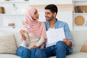 Happy arab couple planning pregnancy, holding positive pregnancy test and ovulation calendar, sitting on sofa