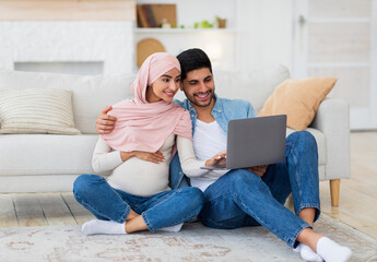 Loving pregnant arab spouses using laptop, sitting on floor at living room, watching baby room interior, free space