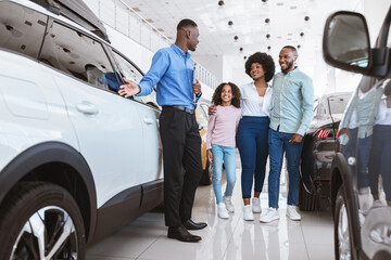 Young black family having conversation with confident sales agent about buying new car at auto...