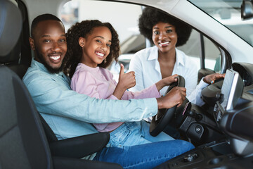 Cute black girl with her parents recommending auto dealership, sitting inside new car, showing thumb up gesture