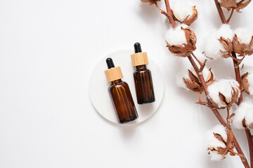 Amber glass dropper bottles with bamboo lid and cotton flowers on white concrete podium. Skincare cosmetic. Beauty concept for face body care. Presentation with daylight.