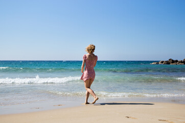 Fototapeta premium unrecognizable young slender woman in a short dress walks along the beach on the coast during the summer holidays. Dreamy, looks at the beautiful sea, fantasies, inspiration, the energy of nature