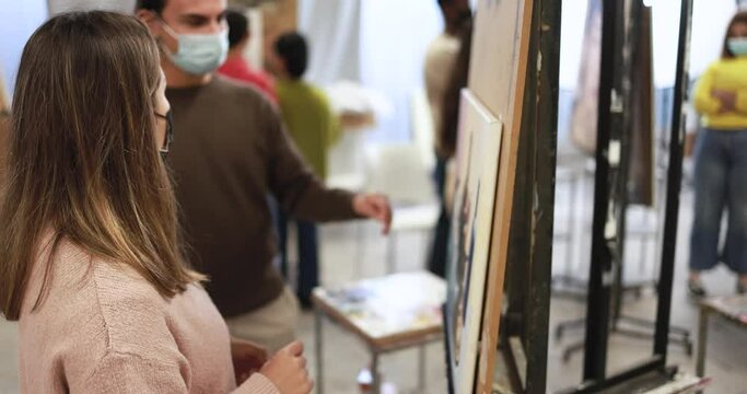 Young students painting inside art room class at college while wearing safety face masks for coronavirus outbreak