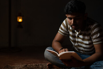 Young student busy reading during night under oil lamp or lantern due to power loss and Poverty -...