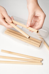 Close-up shot of female hands holding reusable bamboo cocktail straws over the table. There are also a lot of bamboo straws and a transparent case with straws on the white table.