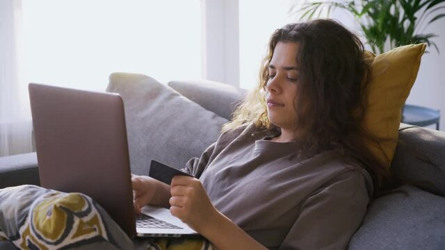  woman relaxing on sofa enters information from credit card typing on laptop keyboard against large window in room in sunny morning, concept of online shopping