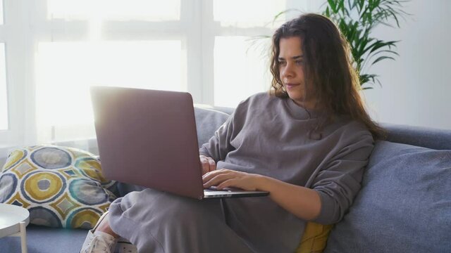 Long haired young woman blogger works on contemporary laptop lying on comfortable grey sofa near large window in sunny morning, freelancer at home