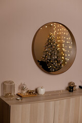 Round mirror with lights and christmas tree reflected in the mirror hanging on beige wall in apartment