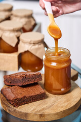 viscous salted caramel flows from a white spoon into a jar. next to it is a piece of chocolate brownie