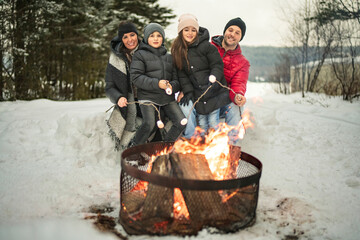 Family winter picnic. Happy friends sit around campfire in forest and fry marshmallows during a...