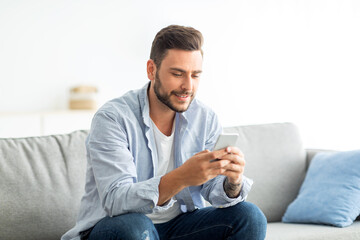 Useful app concept. Millennial man using modern smartphone while sitting on sofa in living room,...
