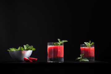 Dos vasos de refrescante jugo de fresa triturado decorado con una rama de menta y fresas cortadas sobre un fondo negro	