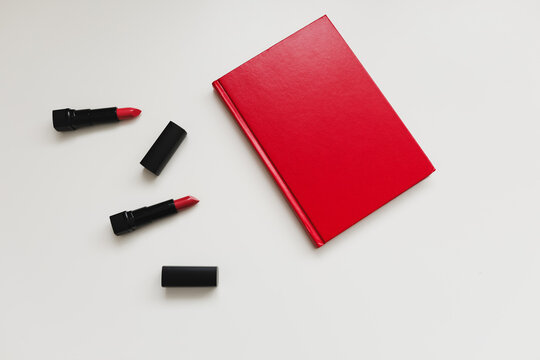 Feminine desk with cosmetics, lipstick and red book or diary on white background. Flat lay, top view.