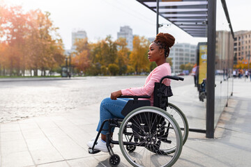 Black paraplegic woman in wheelchair waiting on bus stop, cannot board vehicle suitable for handicapped persons - obrazy, fototapety, plakaty
