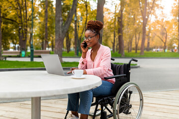 Disabled black lady in wheelchair using laptop, having conversation on smartphone at outdoor cafe...
