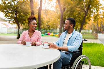 Disabled black guy in wheelchair having date with woman in outdoor cafe, drinking coffee, enjoying talk at autumn park