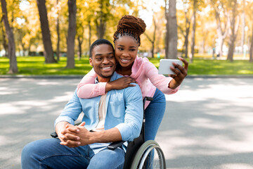 Affectionate black woman and her impaired boyfriend in wheelchair taking selfie together at autumn...
