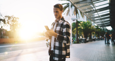 Serious caucasian millennial male traveler watching video via smartphone and earphones while spending time on street in city, hipster guy downloading music to mobile phone playlist on urban setting