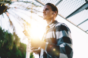 Handsome millennial male in earphones holding mobile phone listening playlist songs via accessory outdoors, 20s man enjoying online audiobook spending free time on street satisfied with 4G connection