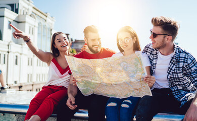 Group of cheerful tourists using travel map for search direction route and explore city during summer vacations, happy Caucasian hipster guys with orientation paper discussing funny journey trip
