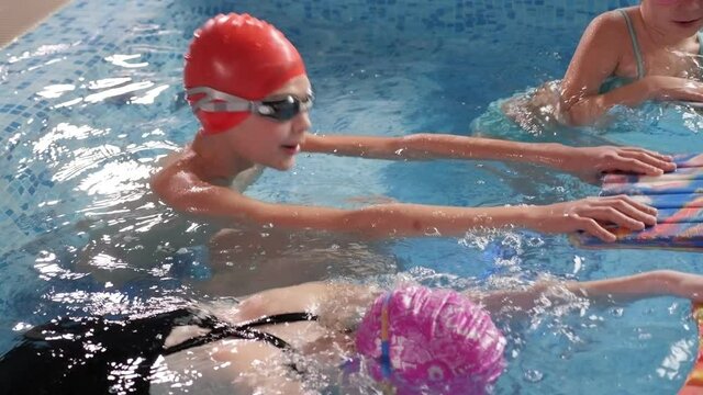 A group of children in swimming caps at a swimming lesson in an indoor pool, they swim with the help of foam boards for swimming.